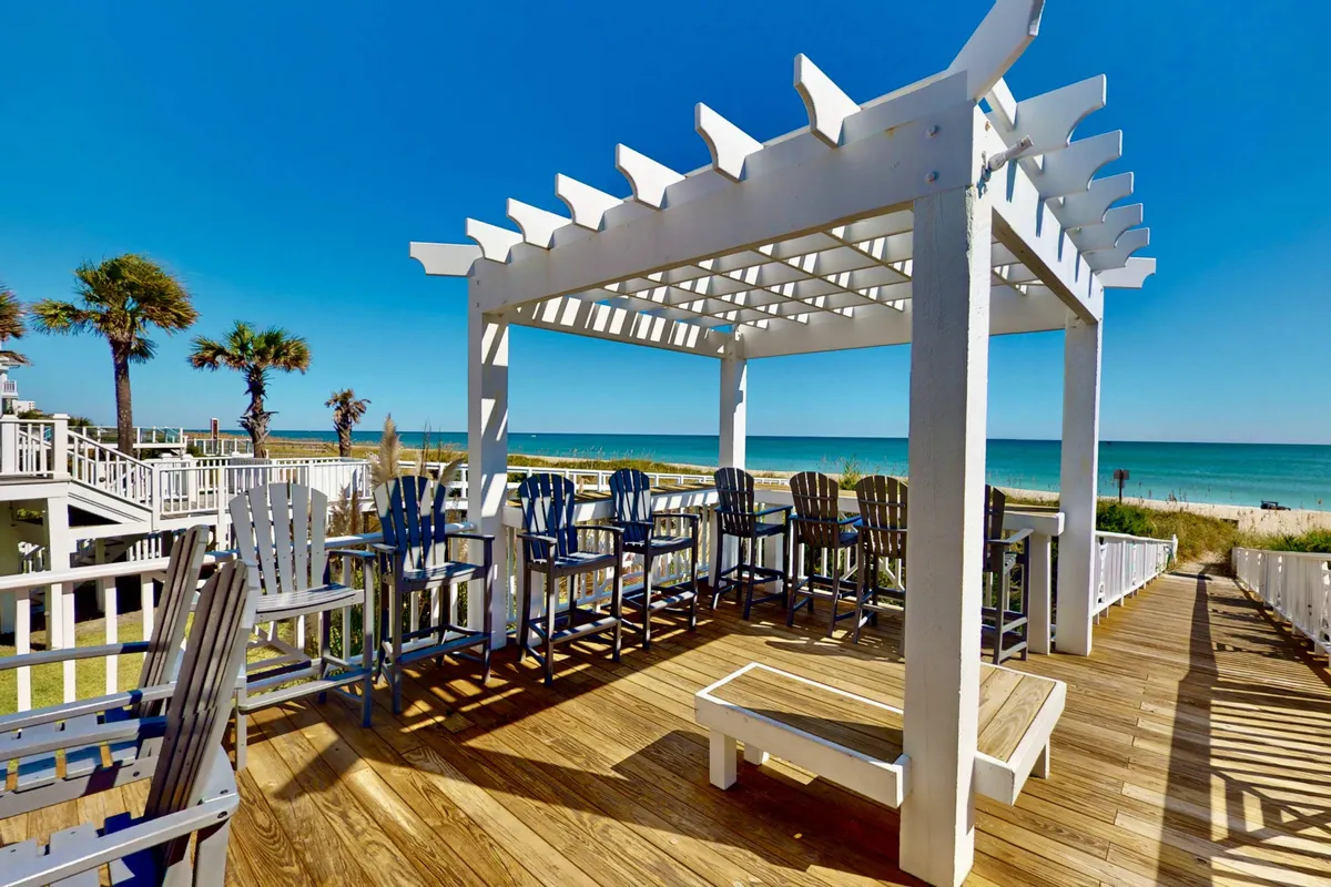 A serene exterior shot featuring a pergola with comfortable outdoor seating, all set against the backdrop of an ocean view. The area is designed for relaxation and socializing, inviting guests to lounge with drinks, enjoy sunset cocktails, or have an afternoon barbecue. Ample natural light and a cheerful mood set the scene for outdoor enjoyment.