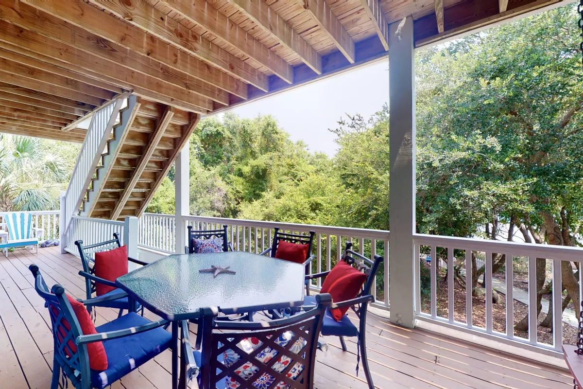 A cozy outdoor deck setup with a glass table featuring a starfish centerpiece, colorful cushioned chairs, and a staircase leading to an upper level. The scene is surrounded by lush greenery, inviting guests to relax or dine al fresco.