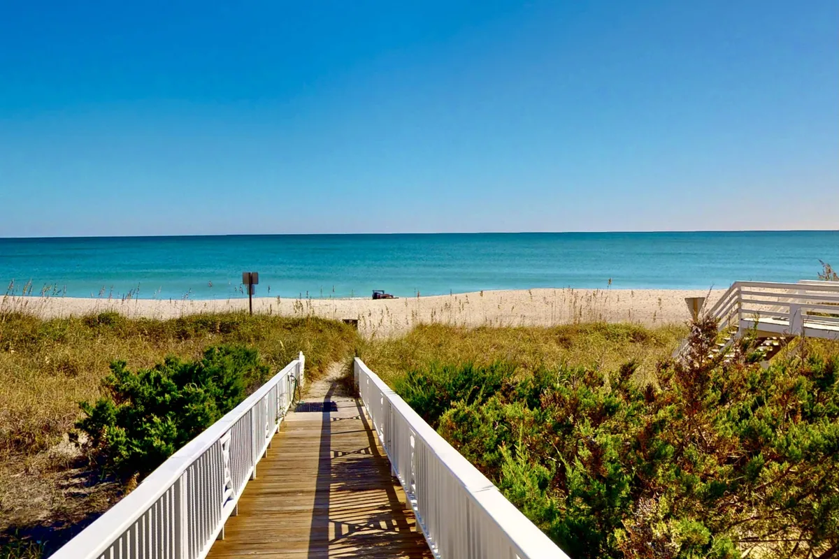 A wooden walkway leads over sand dunes to a pristine beach with gentle waves and a clear blue sky. The scene suggests easy access to the ocean, with ample space for walking, playing, or relaxing by the shore.