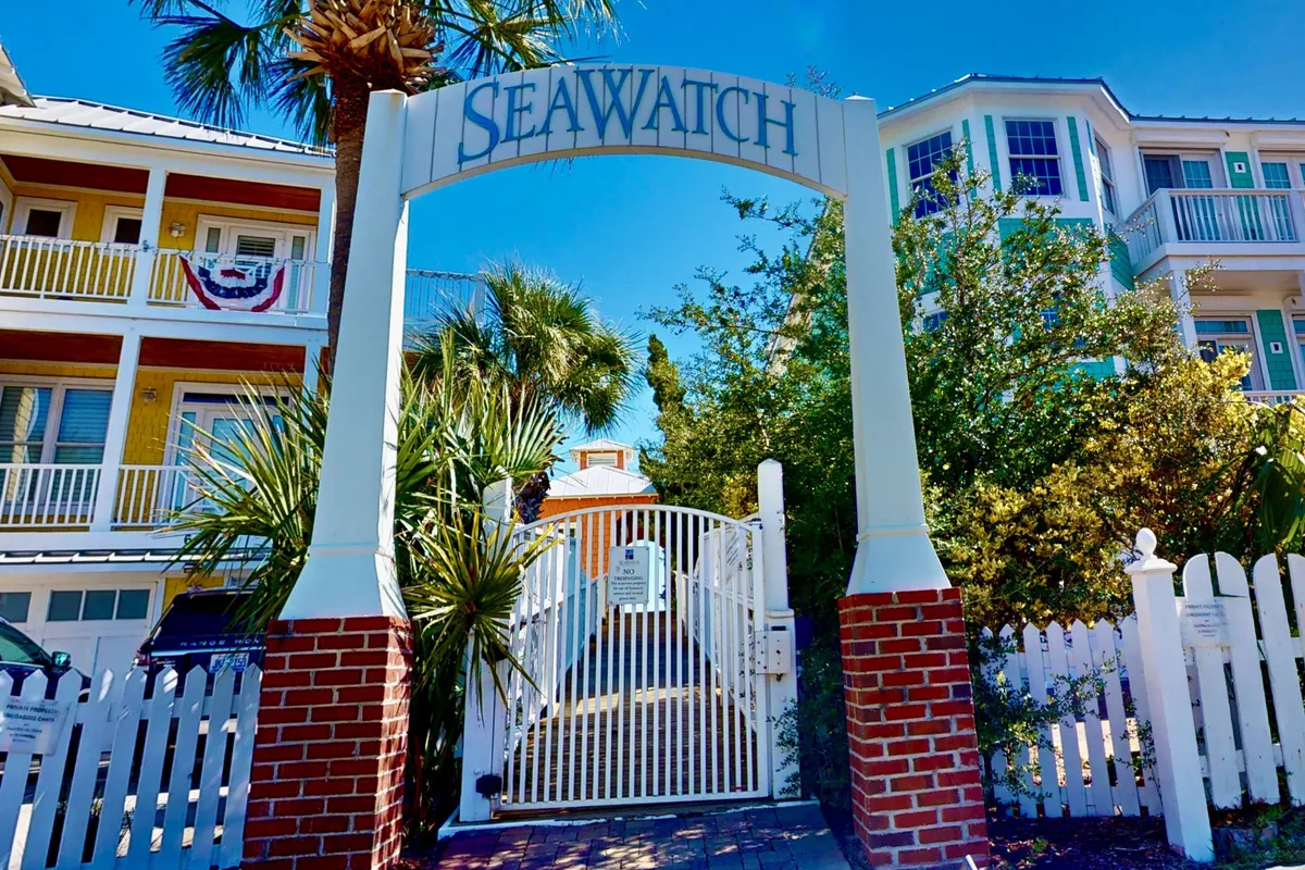This exterior shot captures the welcoming archway with the 'SEA WATCH' sign, white picket fence, palm trees, and a glimpse of colorful buildings beyond. The scene feels vibrant and inviting, with a landscaped garden area perfect for strolling or relaxing.