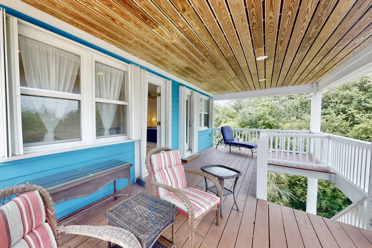 A relaxed porch scene with blue exterior walls, striped chairs, a wooden ceiling, and views of lush greenery. The seating arrangement encourages conversation or quiet moments with coffee, offering a comfortable transition between private and social spaces.