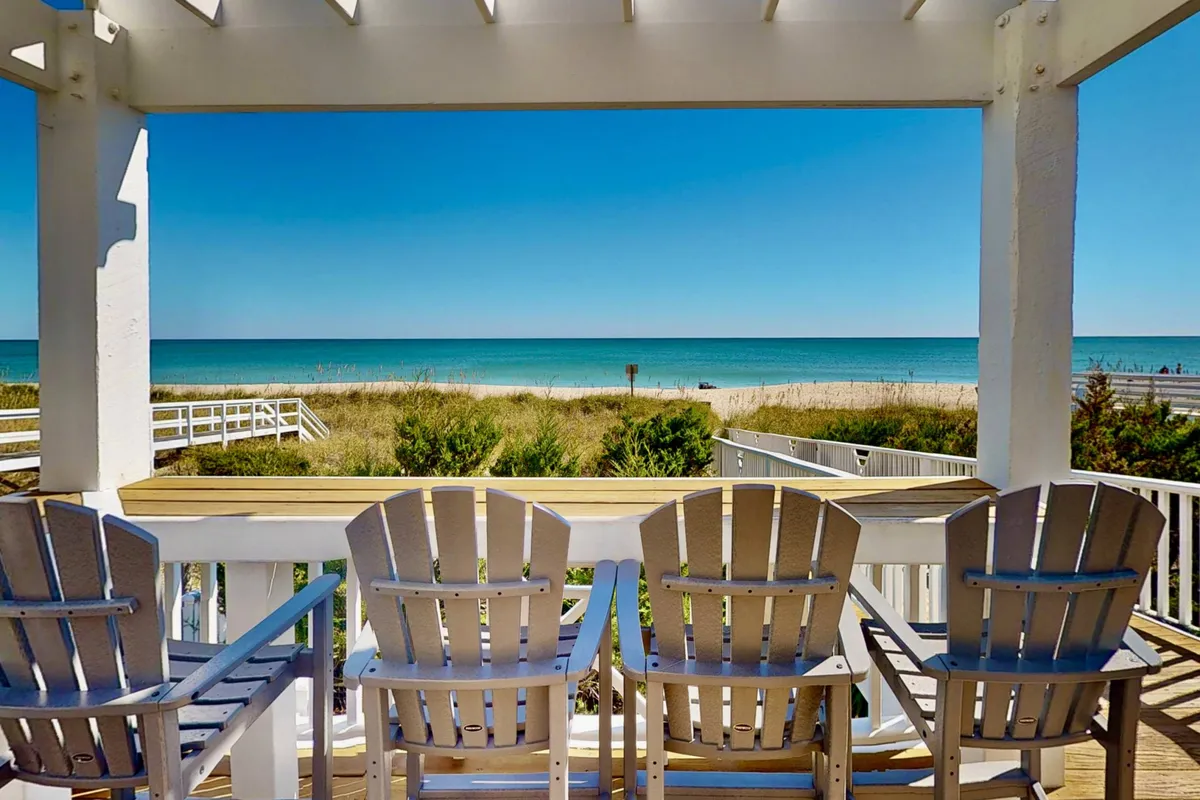 A line of Adirondack chairs faces sweeping ocean views over sand dunes and under a vast blue sky. The peaceful scene is perfect for enjoying sunrises, sunsets, or quiet moments with friends.
