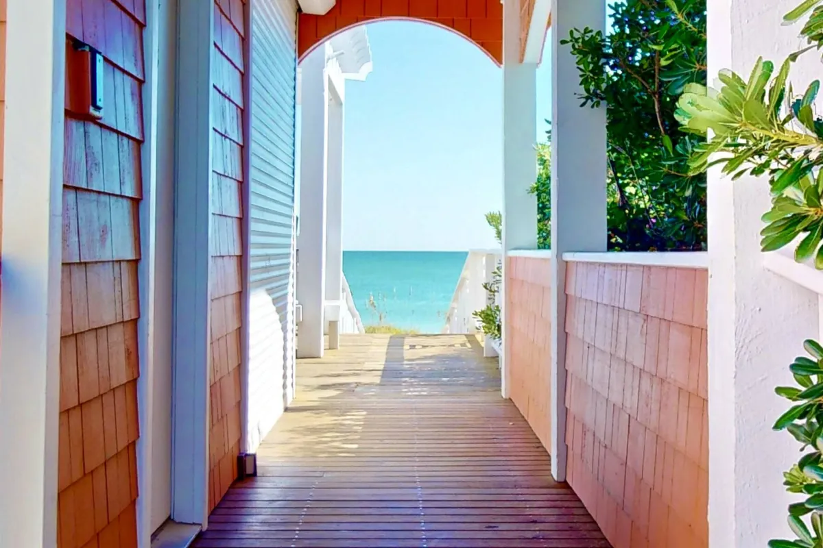 A serene exterior featuring a wooden walkway leading to an ocean view, framed by an arched entrance and green foliage. The scene suggests easy movement and accessibility, inviting groups to stroll, enjoy the breeze, or gather for conversations with a beautiful natural backdrop.