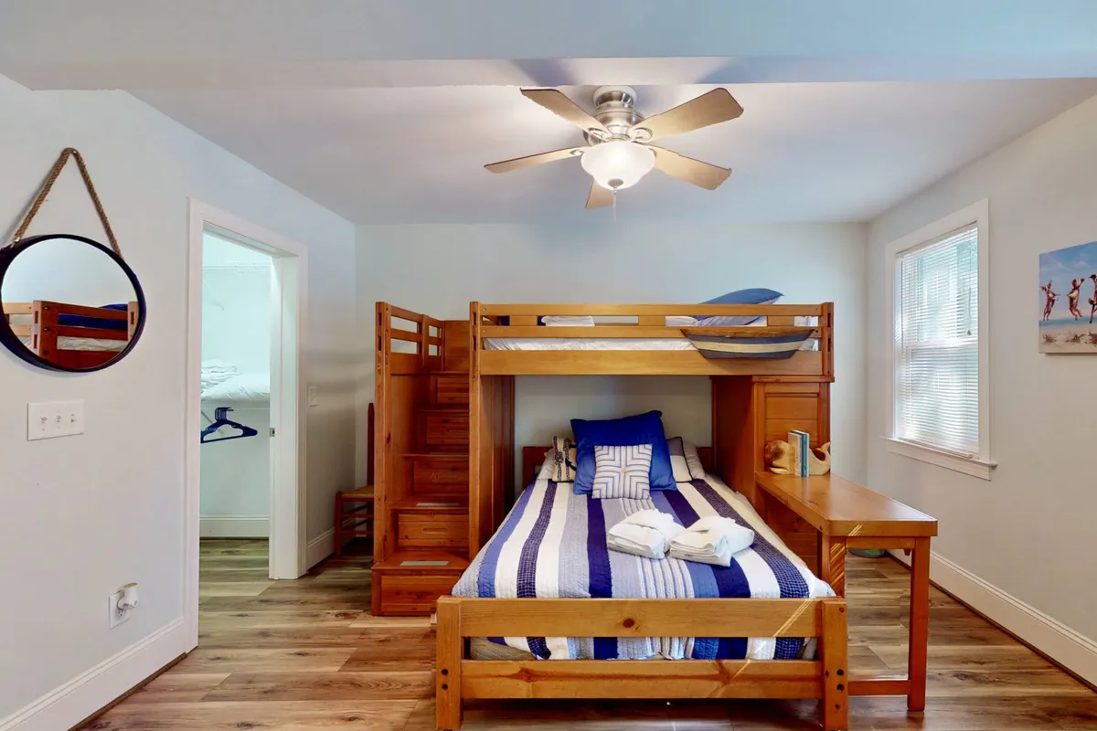 A playful bunk room featuring striped bedding on a bunk bed, wooden furniture, a ceiling fan, and cheerful wall art. The room radiates a fun, cozy energy perfect for children or group gatherings.