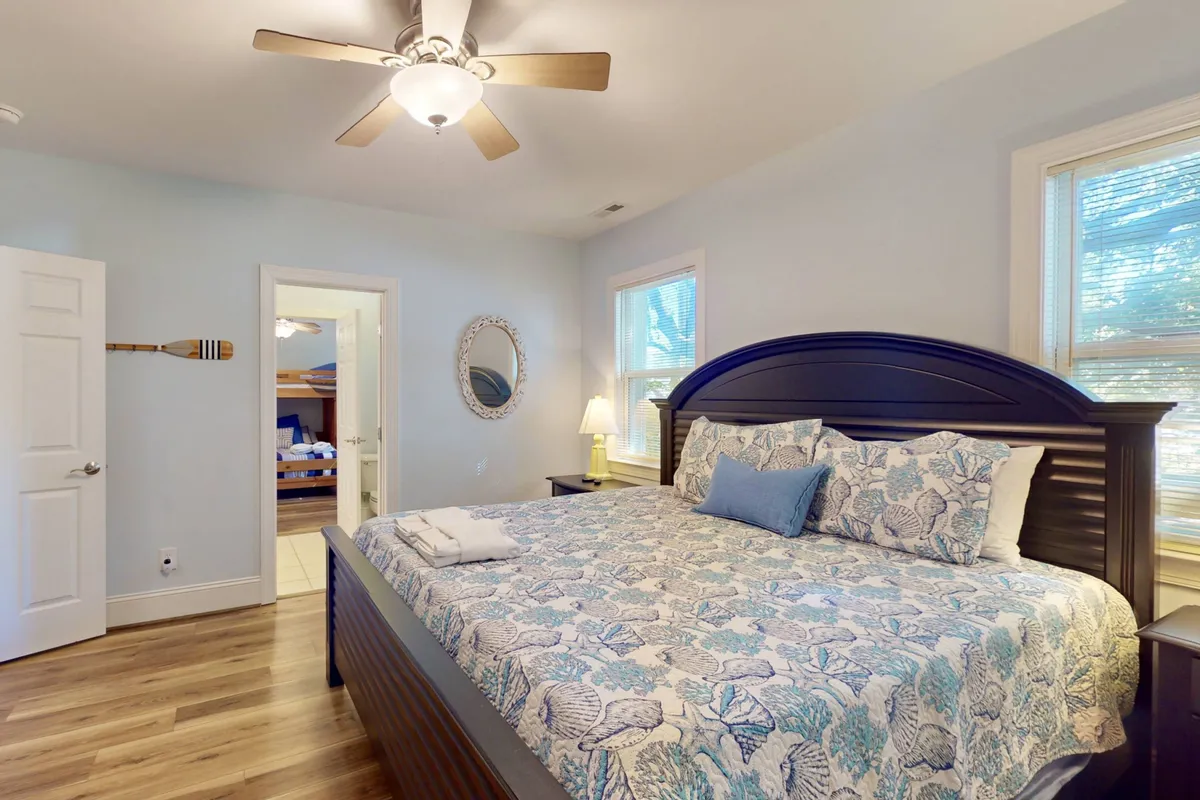A king-sized bed with ocean-themed bedding sits in a calming bedroom flooded with natural light from a large window. A decorative mirror and a wooden paddle on the wall add coastal charm, creating an inviting atmosphere for relaxation.