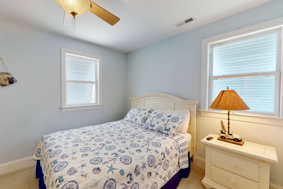 A queen bedroom features blue and white patterned bedding, a bedside lamp, and a window letting in soft sunlight. The space feels tranquil and comfortable, ideal for restful nights and quiet moments.
