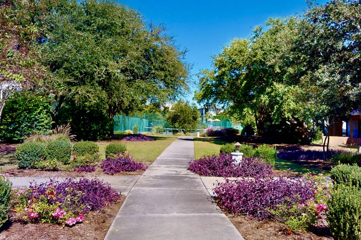 A serene pathway winds through beautifully landscaped flower beds and mature trees, offering a tranquil, shaded environment ideal for leisurely walks. The greenery and blooming plants evoke a peaceful, natural setting, perfect for morning strolls with pets.