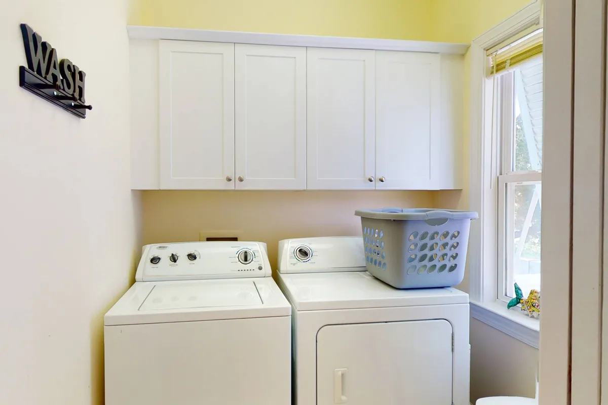 A sunlit laundry area featuring a modern washing machine and dryer, with a laundry basket nearby and a window allowing natural light to brighten the space. The arrangement is neat and functional, communicating ease and comfort for guests handling everyday chores.
