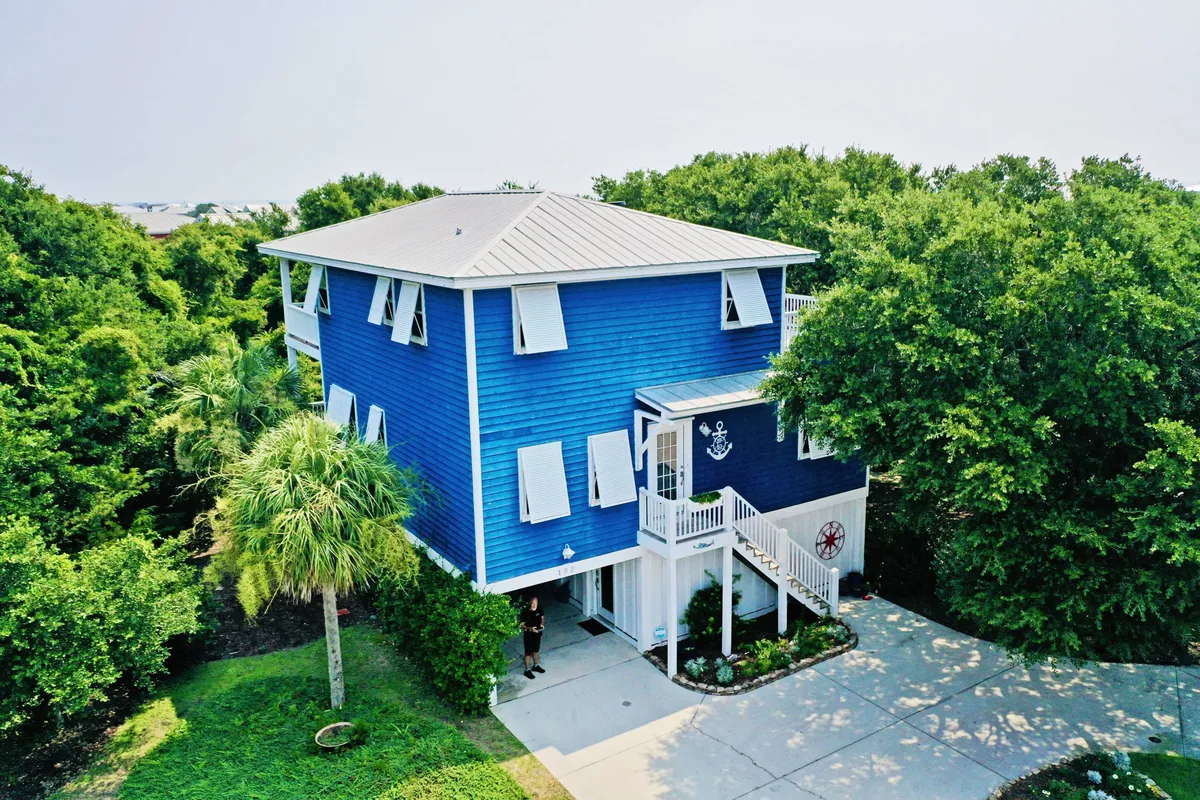 The image showcases a charming blue single-family house nestled among lush greenery, with a spacious front porch adorned with nautical decorations. The inviting setting hints at tranquil moments on the porch, surrounded by nature, and the upscale neighborhood of SeaWatch in Kure Beach.